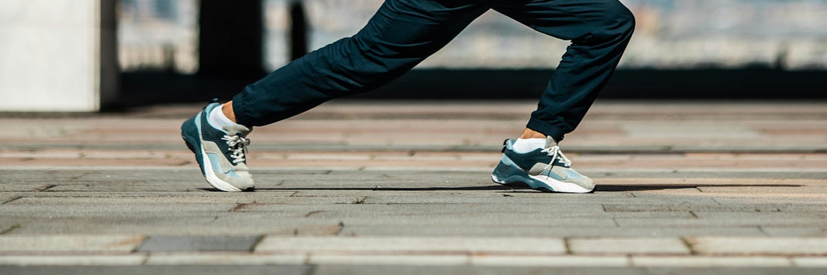 Fotografie vom Unterkörper eines Mannes der eine Dehnübung ausführt und dabei eine blaue Hose sowie Turnschuhe trägt 